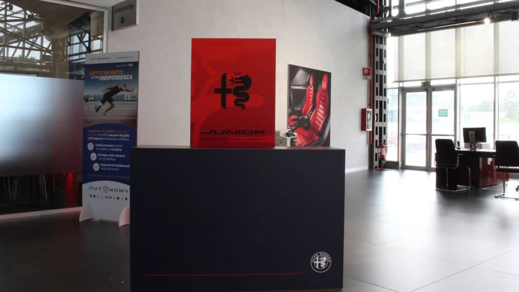 Uno stand espositivo nell'atrio di un moderno ufficio espone materiale promozionale del settore automobilistico, tra cui un cartello rosso con il logo di un'auto e due striscioni informativi sullo sfondo.